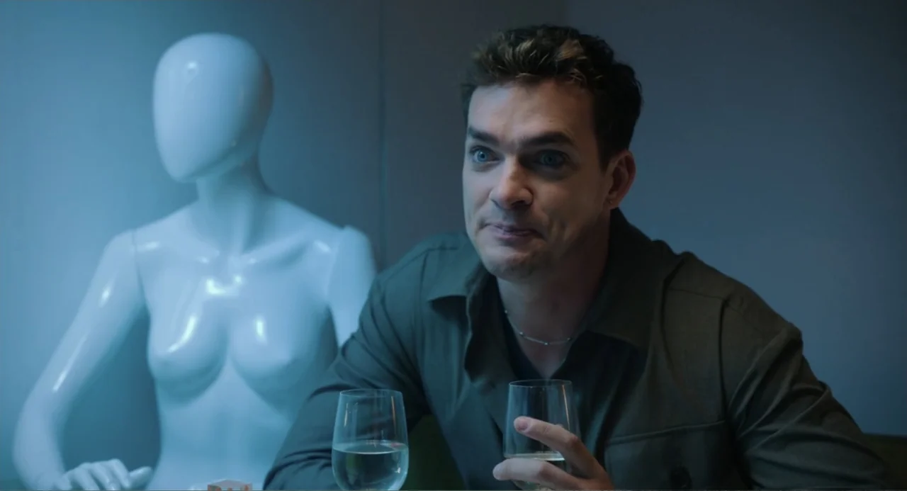 Picture This (2008) movie still: medium — A man sits at a table in a dimly lit room with a mannequin behind him. He holds a glass of water while…