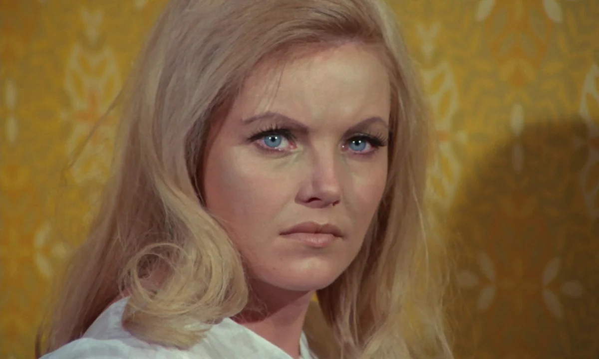 Vampyros Lesbos (1971) movie still: closeup — A close-up shot of a woman against a yellow patterned wallpaper background. The subject maintains…