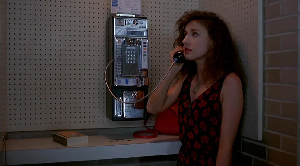 Mystery Train (1989) movie still: medium — A corner of a room features a wall-mounted public coin-operated telephone and pegboard wall. The woman…