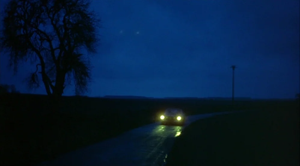 Un Flic (1972) movie still: wide — A dark rural road stretches through a flat landscape under a deep blue night sky. A car moves forward on the wet…