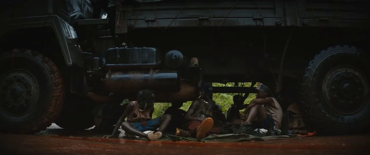 Beasts of No Nation (2015) movie still: wide — A group of teenagers sits on a groundsheet beneath the dark undercarriage of a truck. The ground is…