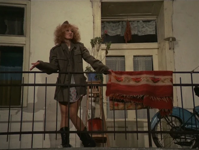 WR: Mysteries of the Organism (1971) movie still: fullbody — A woman stands on a balcony against a white wall with an open window behind. A…