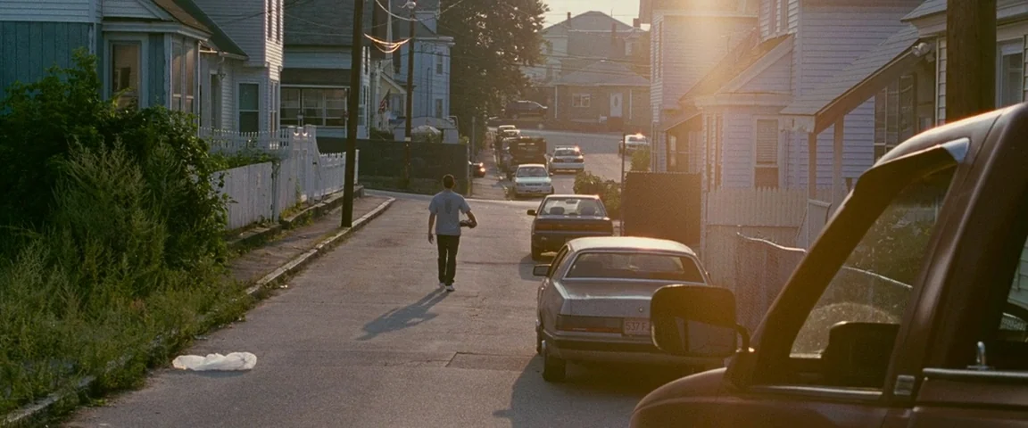 The Fighter (2010) movie still: wide — A residential street at sunset with wooden houses and parked cars lining the pavement. A lone man in a…