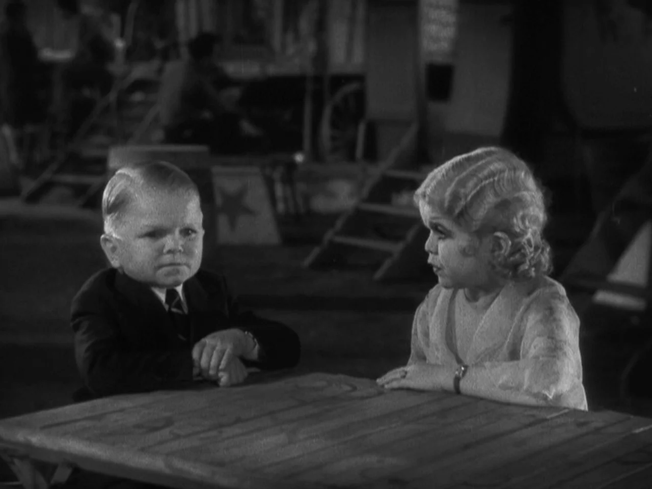 Freaks (1932) movie still: medium — A wooden table is positioned in an outdoor carnival grounds area with equipment in background. A man and woman…
