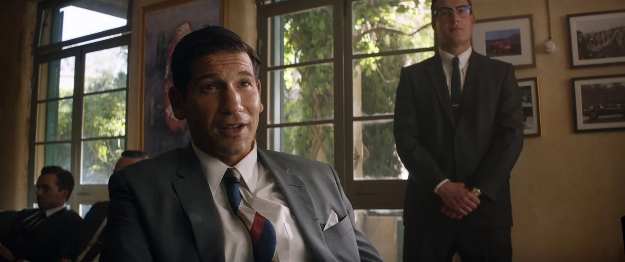 Ford V Ferrari (2019) movie still: medium — A sunlit office features large glass doors and framed vintage car racing prints. One man sits in the…