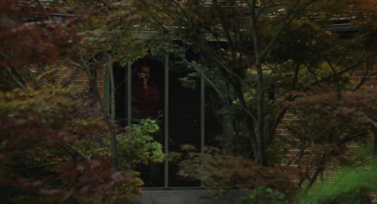 The Nest (2020) movie still: wide — A brick-walled building is viewed through a dense frame of reddish-green Japanese maple leaves. A man stands…