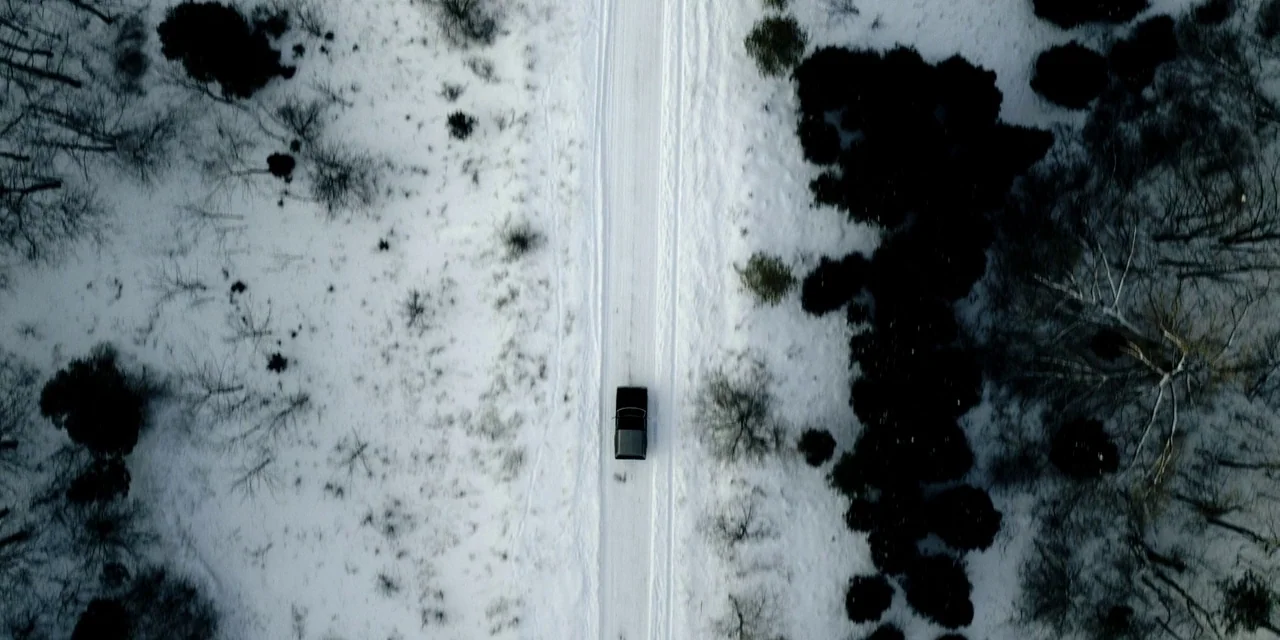 Polar (2019) movie still: establishing — A dark vehicle travels along a narrow, snowy track flanked by bare, wintry trees. The overhead perspective…