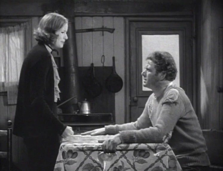 Anna Christie (1930) movie still: medium — A rustic dining room interior features wooden walls, a stove, and hanging cookware. A woman standing by a…