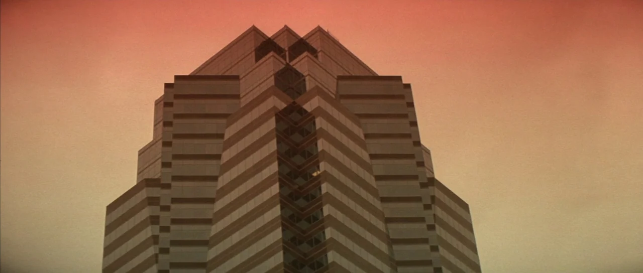 Die Hard (1988) movie still: establishing — A tiered glass and concrete corporate skyscraper stands against a vibrant orange evening sky. The…