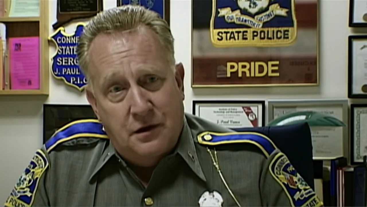 The Cheshire Murders (2013) movie still: closeup — A police officer sits in an office with various framed plaques on the walls. The man wears a…