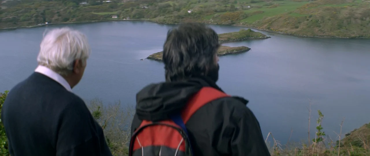 Silence (2012) movie still: medium — Two men stand on a cliff edge overlooking a tranquil, dark blue coastal inlet. An island sits in the middle of…