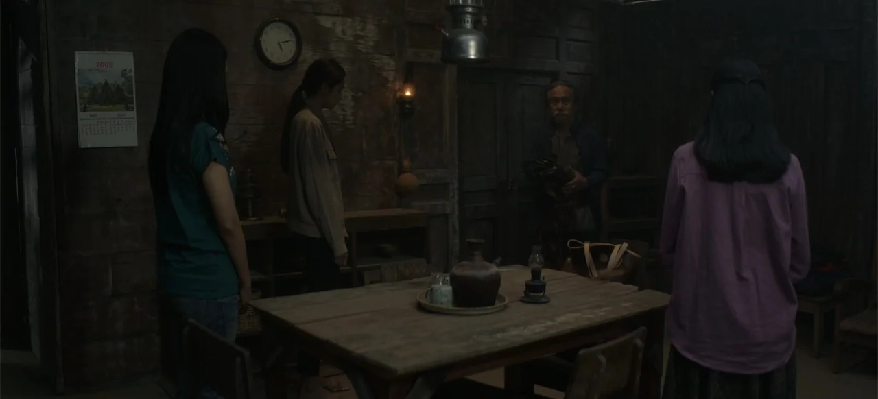 A Thousand Days (2023) movie still: medium — A dark, rustic interior features wooden plank walls and a central dining table. Three women stand…