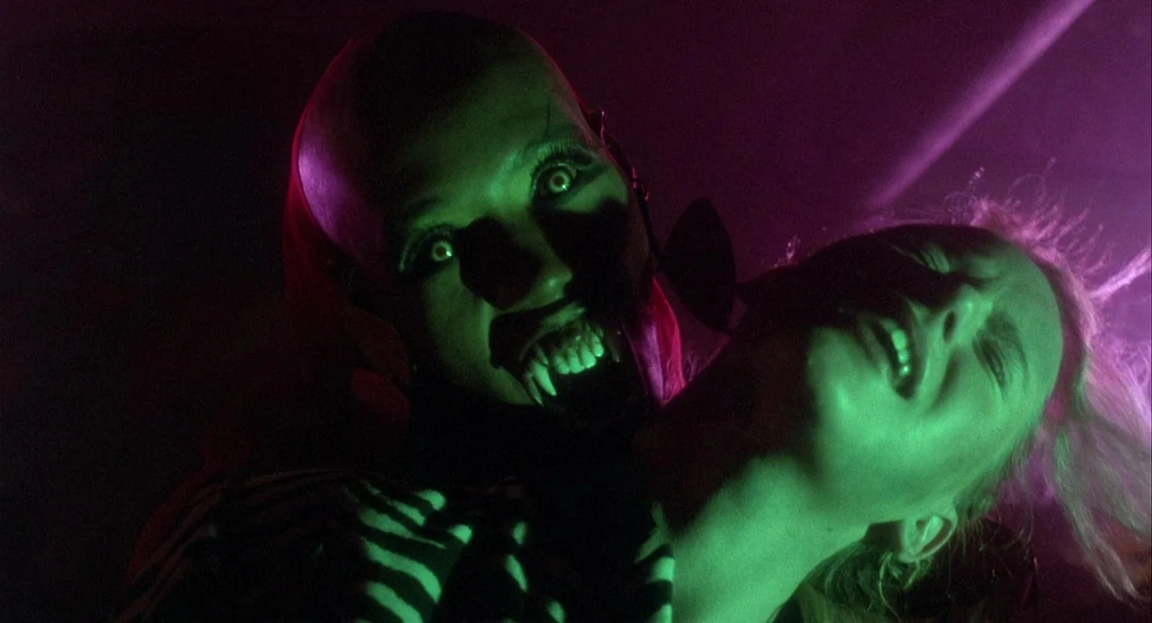 Vamp (1986) movie still: closeup — A dimly lit space features strong green and magenta light sources on two people. A bald, fanged figure grips a…