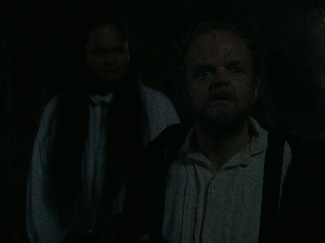 First Cow (2019) movie still: medium — A dimly lit nighttime environment with dense, unidentifiable surroundings obscured by deep shadows. Two men…