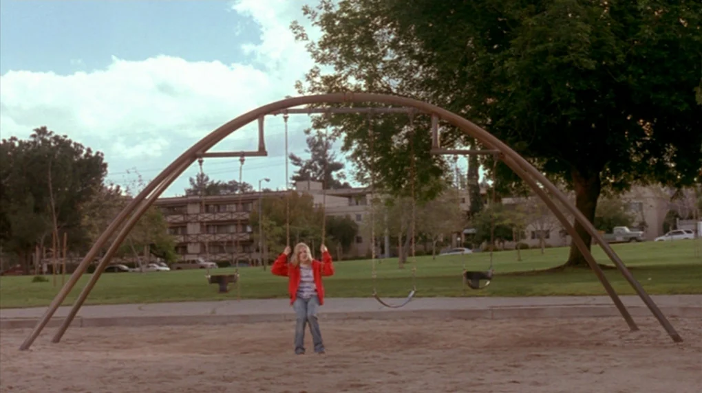 Smiley Face (2007) movie still: wide — A public playground features a large arched metal swing set on sandy ground. A teenage girl stands in front…