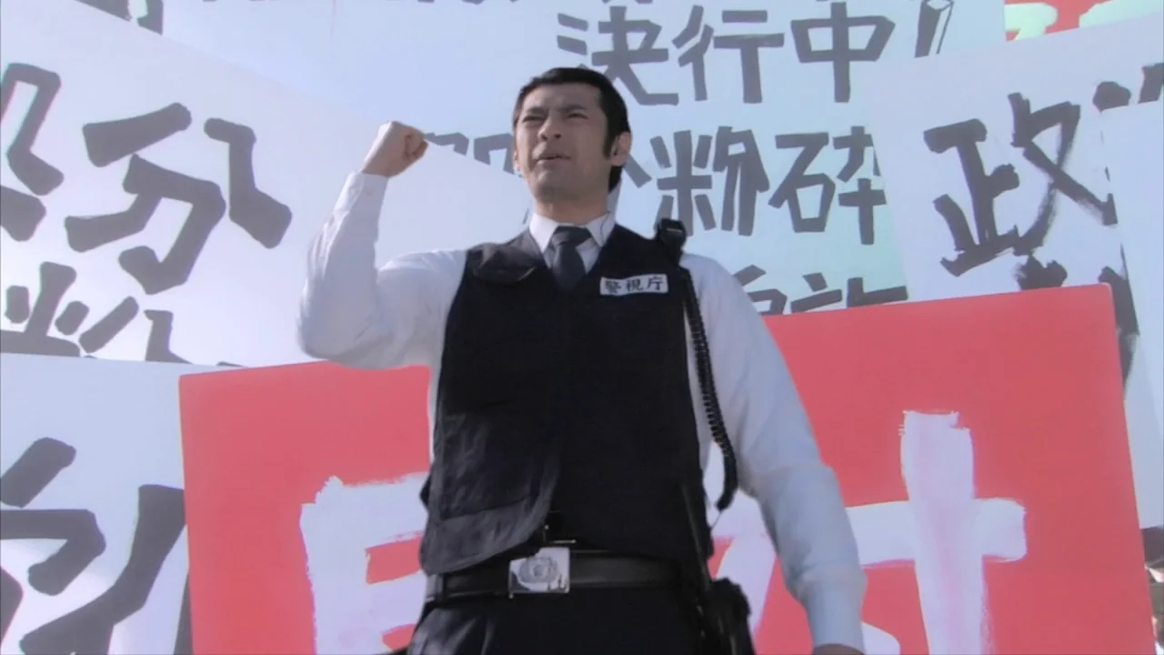 Tokyo Gore Police (2008) movie still: medium — A man stands outdoors in front of multiple large protest placards with Japanese text. The individual…