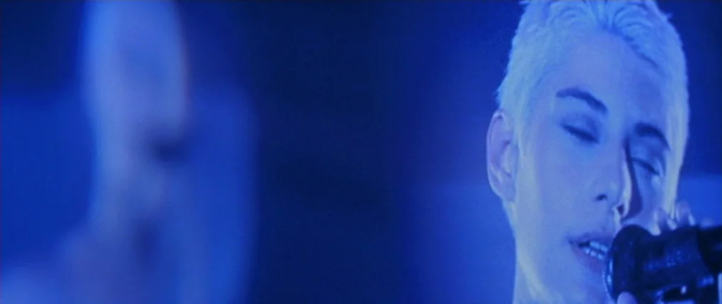 I Want You (1998) movie still: closeup — A person with short bleached hair performs on a dimly lit dark stage. The individual holds a microphone and…