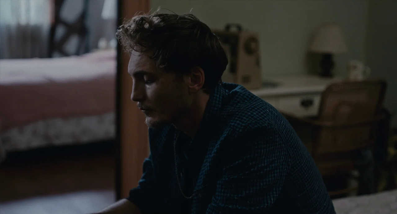 Lingua Franca (2019) movie still: medium — A furnished bedroom features a wooden desk in the background and an unmade bed. A young man sits in the…
