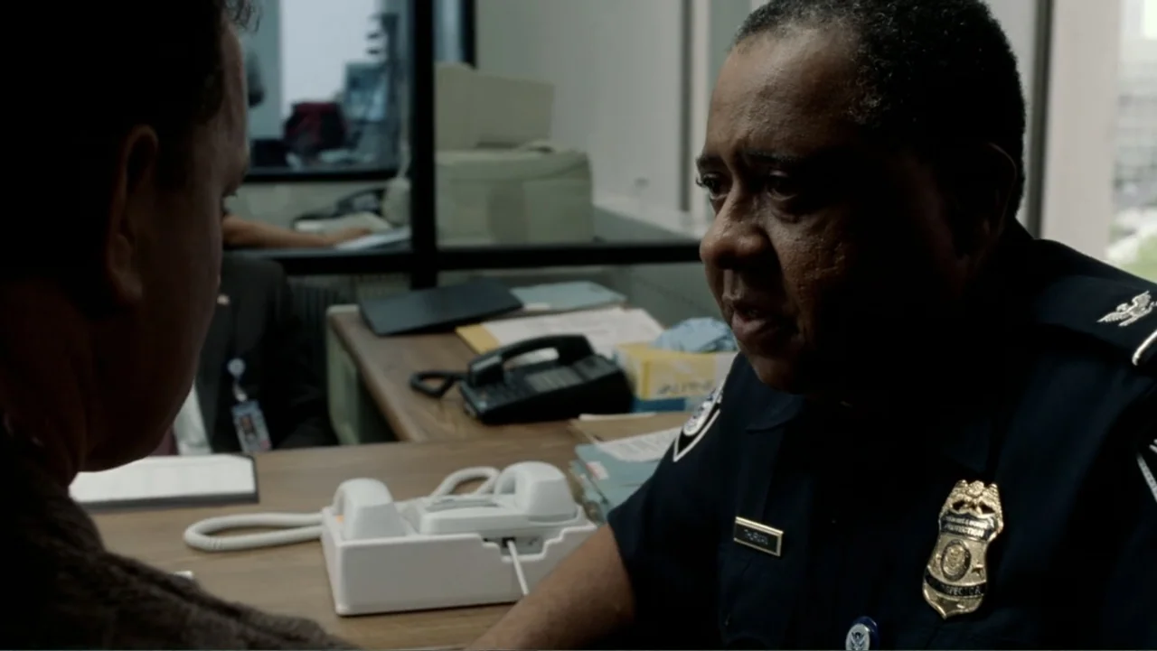 The Terminal (2004) movie still: medium — A cramped government office features a wooden desk with a telephone and file folders. A uniformed…