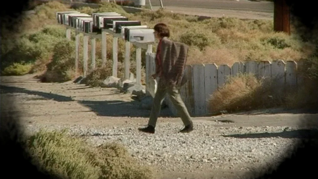 Land of Plenty (2004) movie still: fullbody — A dry, sun-baked roadside area features a line of metal mailboxes on posts. A woman walks horizontally…
