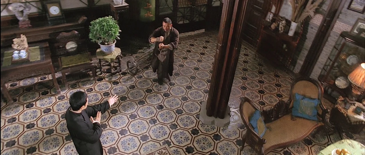 Ip Man (2008) movie still: wide — A spacious room features ornate patterned floor tiles and wooden architectural support columns. Two men wearing…