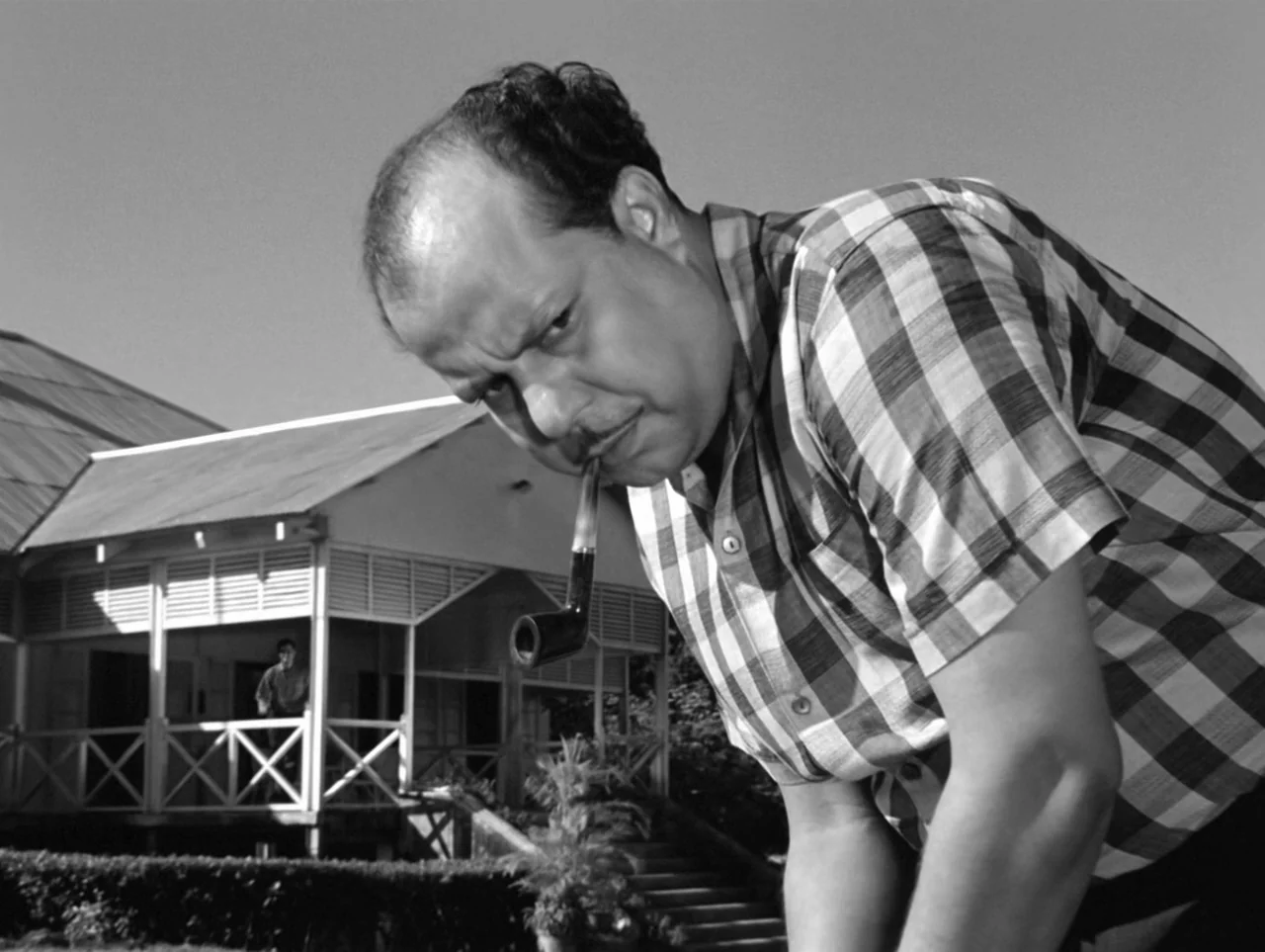 The Coward (1965) movie still: medium — A man in a plaid shirt stands in the foreground in front of a house. A second person is visible standing on…