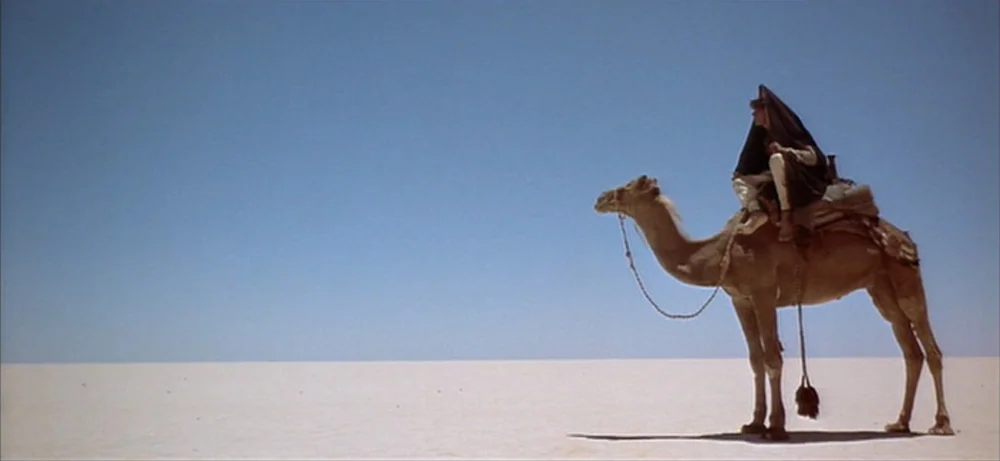 Lawrence Of Arabia movie still: wide — A vast, flat desert landscape stretches toward a clear blue sky. A single man sits atop a camel positioned on…