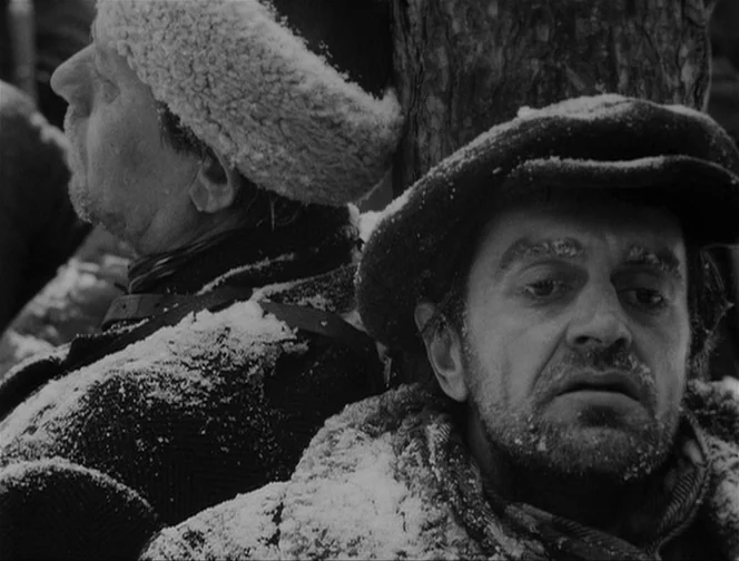 The Ascent (1977) movie still: medium — A snow-covered woodland setting with two men resting against the rough bark of a tree. They wear thick…