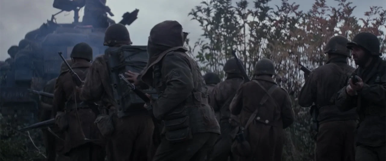 Fury (2014) movie still: wide — A squad of soldiers in 1940s combat gear moves through dense field vegetation. They advance cautiously alongside a…