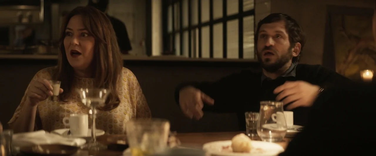 Voy a pasármelo bien (2022) movie still: medium — A dimly lit restaurant table is cluttered with plates, glasses, and small coffee cups. A man and a…