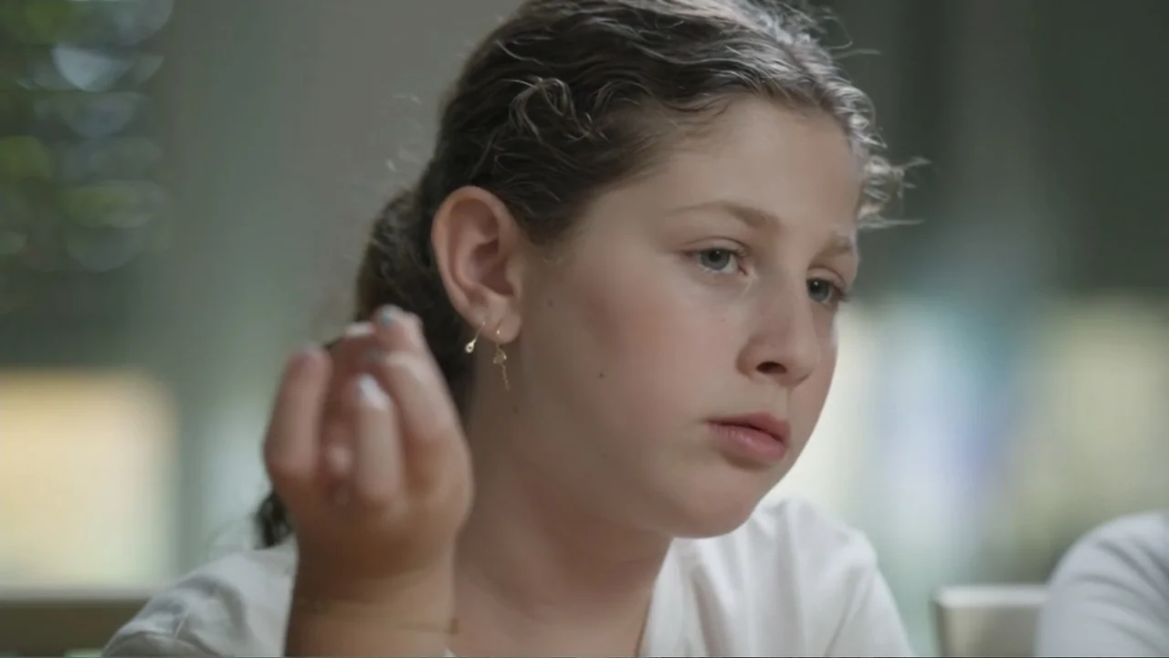 The Children of October 7 (2025) movie still: closeup — A light-toned interior space with soft diffused light levels. A young girl with wavy brown…