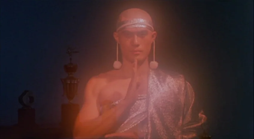 Boxer’s Omen (Mo) (1983) movie still: medium — A dark, dimly lit interior space features a silver-clad man positioned centrally. The man performs a…