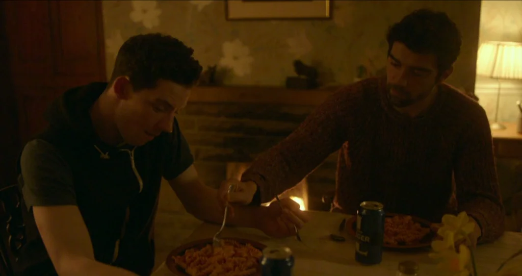 God’s Own Country (2017) movie still: medium — A dimly lit room features a wooden dining table with plates of food. Two men sit across from each…