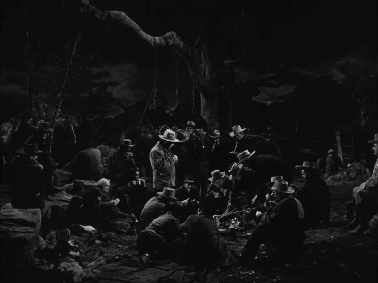 The Ox-Bow Incident (1942) movie still: wide — A group of men sit around a campfire beneath the twisted branches of a tree. Three noose ropes hang…