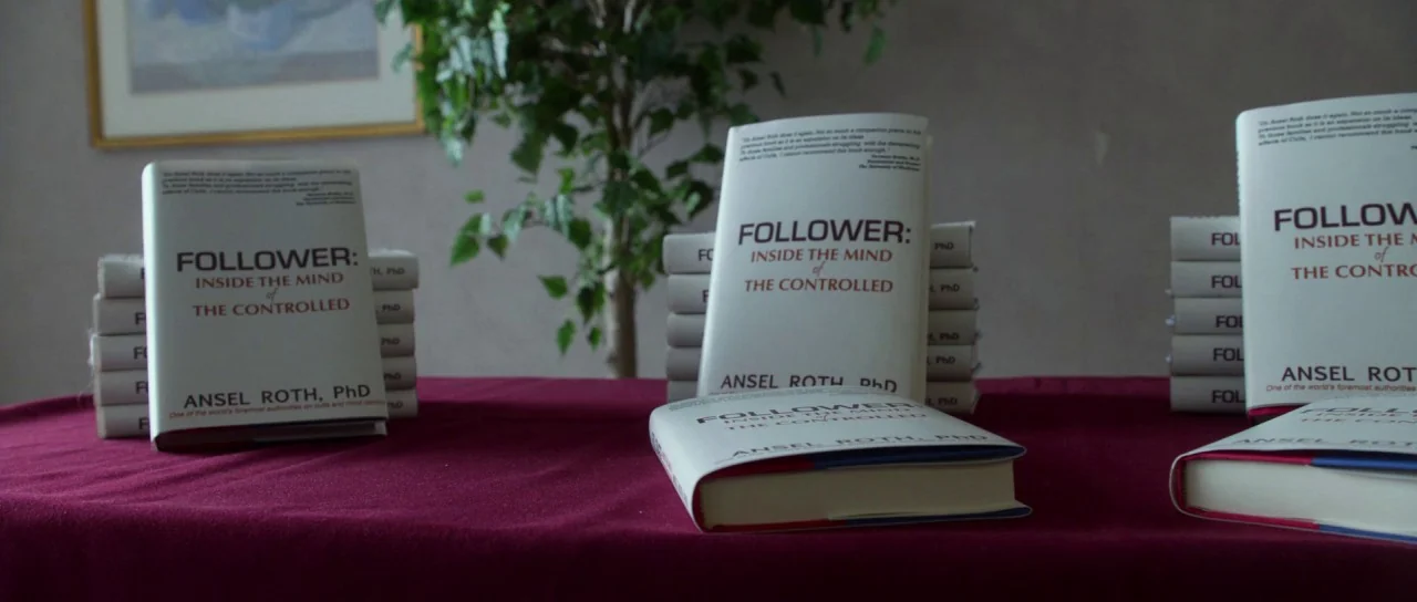 Faults (2014) movie still: medium — A maroon-draped table holds several stacks of books titled Follower Inside the Mind of the Controlled. A small…