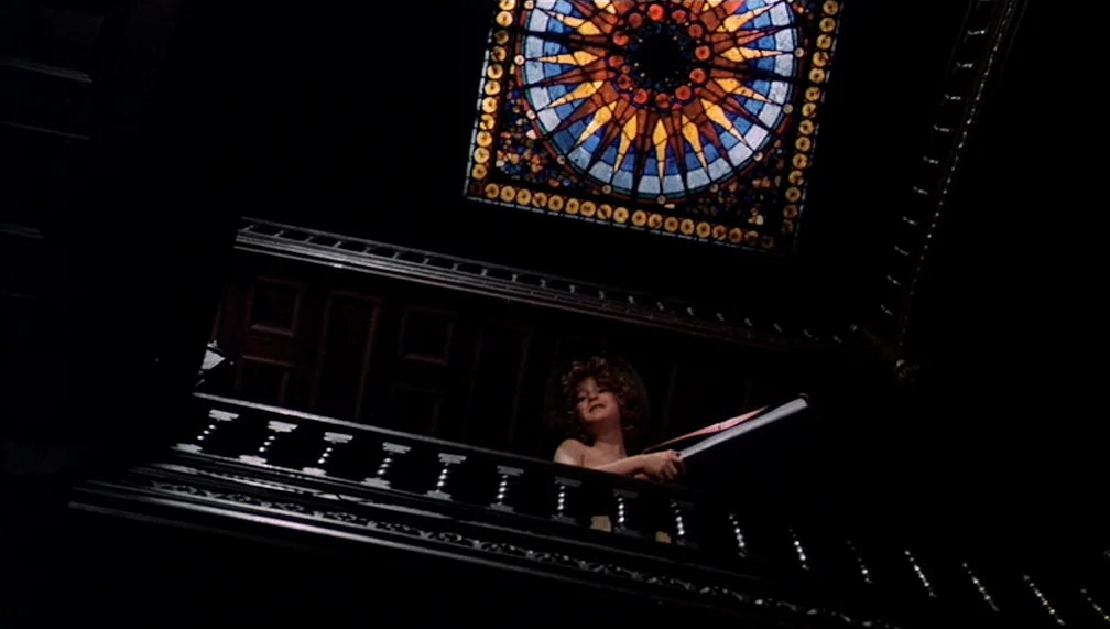 Pretty Baby (1978) movie still: wide — A dark wooden staircase landing features a large circular stained glass window above. A young girl with curly…
