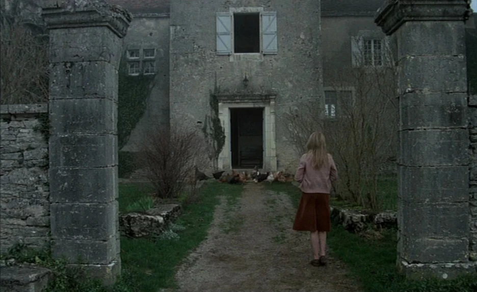 Black Moon (1975) movie still: fullbody — A stone estate features an open doorway flanked by two large, weathered masonry pillars. A young woman…