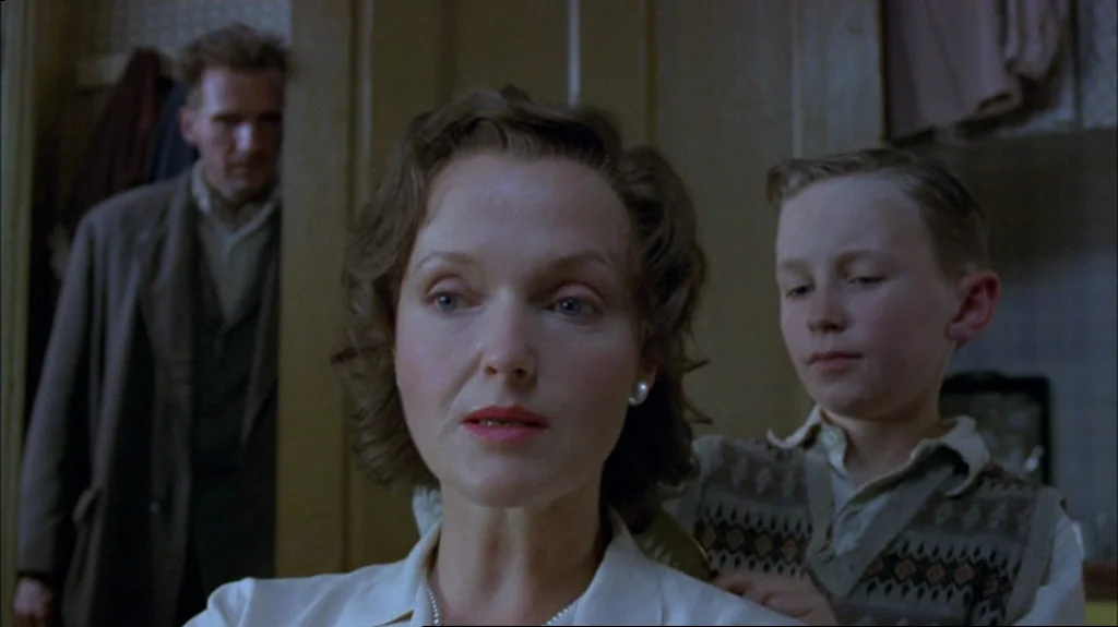 Spider (2002) movie still: medium — An interior space showing a woman, a young boy, and a man in the background. The boy uses a brush to groom the…