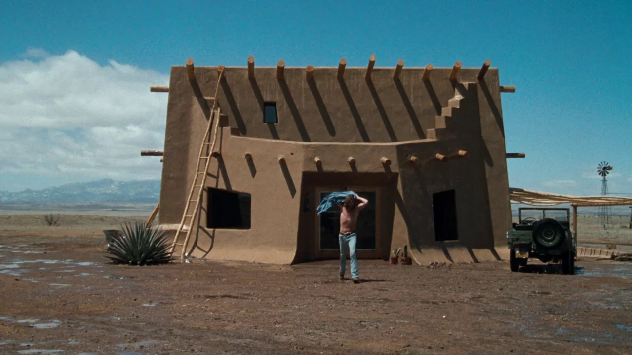 A Star Is Born (1976) movie still: wide — A single-story adobe home stands in a vast, dry desert landscape under a clear sky. A man exits the…