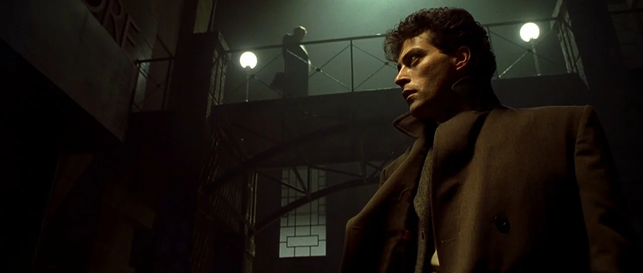 Dark City (1998) movie still: medium — A dark outdoor space features an elevated metal walkway illuminated by glowing streetlamps. A man in an…