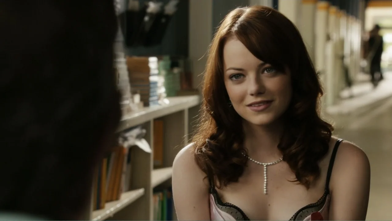 Easy A (2010) movie still: medium — A school hallway interior features lockers along one wall with books visible on shelves. A young woman with…