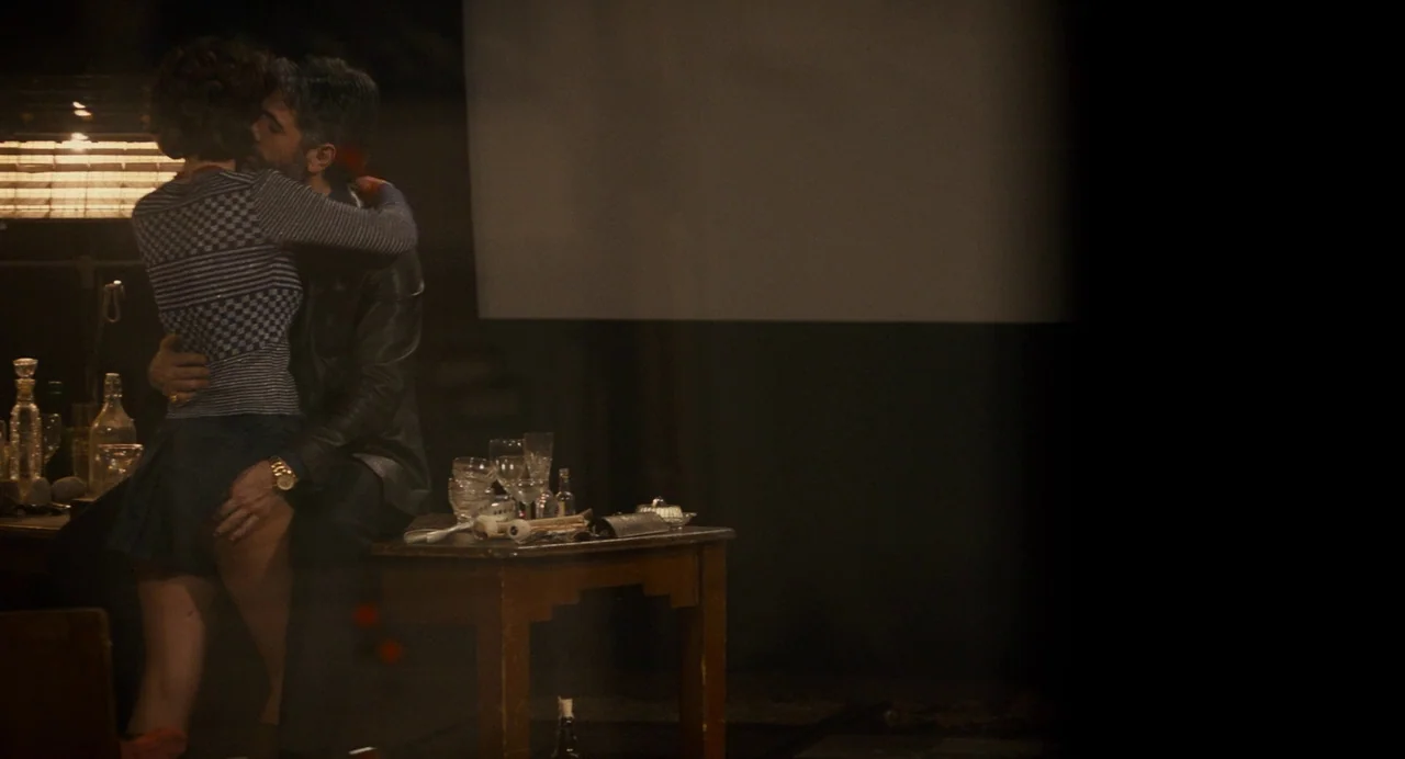 Berberian Sound Studio (2012) movie still: medium — A dimly lit area with a wooden table holding scattered glassware and bottles. Two people embrace…