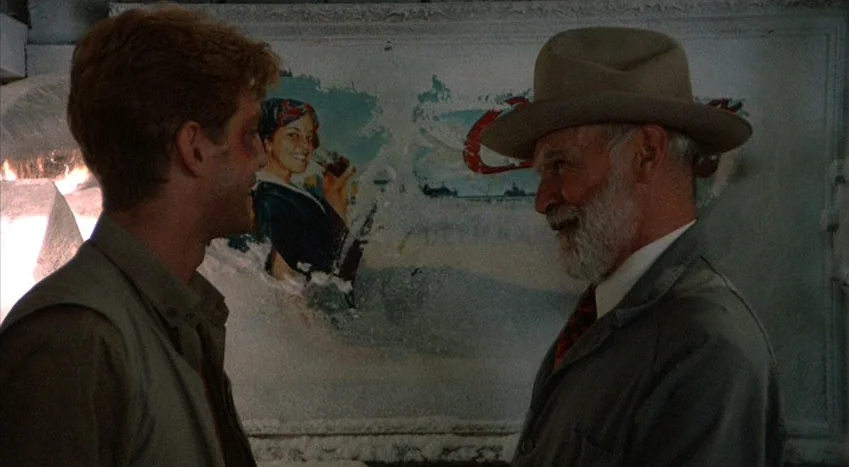 The Coca-Cola Kid (1985) movie still: medium — Two men converse inside a darkened shop space with an old illuminated ice chest. The cooler features…
