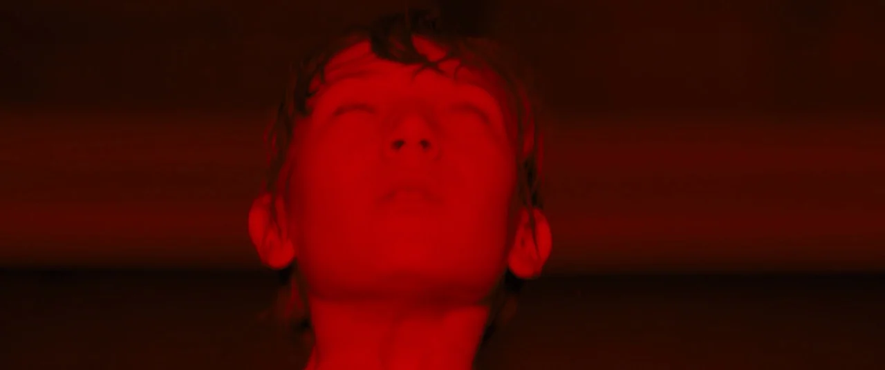 Brightburn (2019) movie still: closeup — A dark space lit only by a strong crimson light source. A young man tilts his head back with his eyes shut…