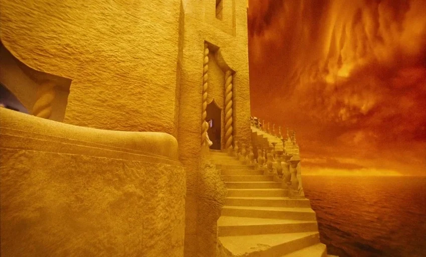 Poor Things (2023) movie still: establishing — A stone palace structure sits beside a dark ocean under a thick orange sky. A curved staircase leads…