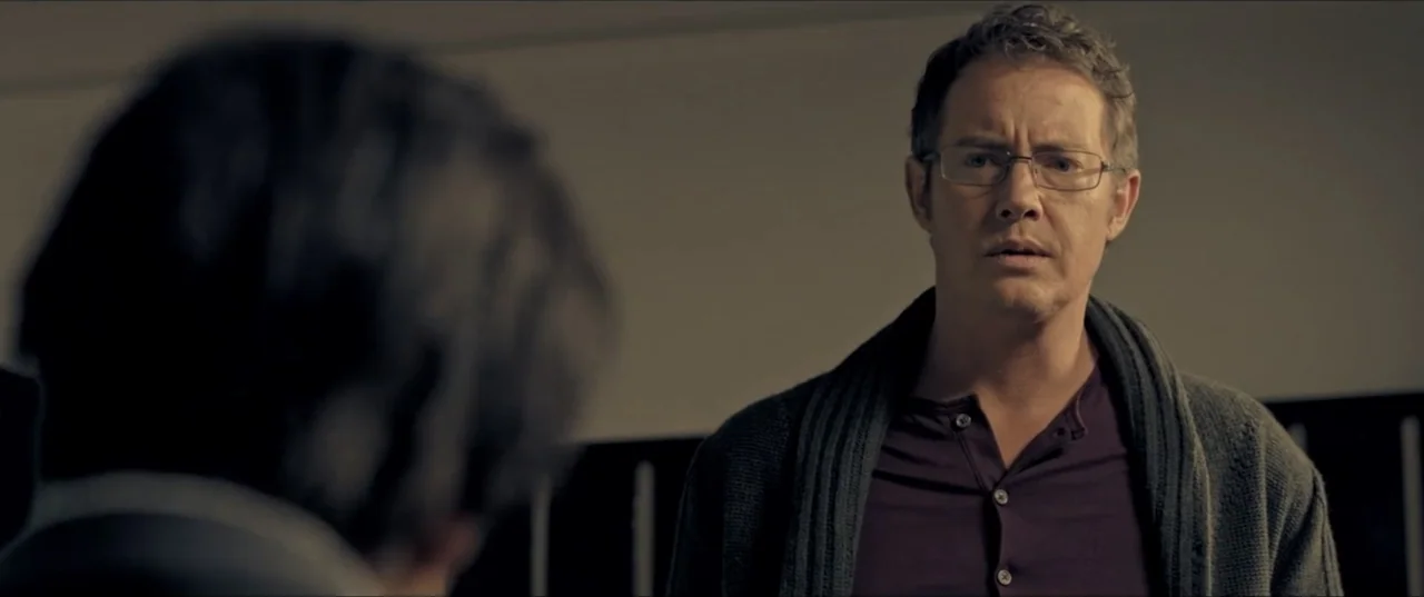 Room 6 (2006) movie still: medium — An interior office space with plain walls and dark vertical structural elements. A man in a cardigan looks…