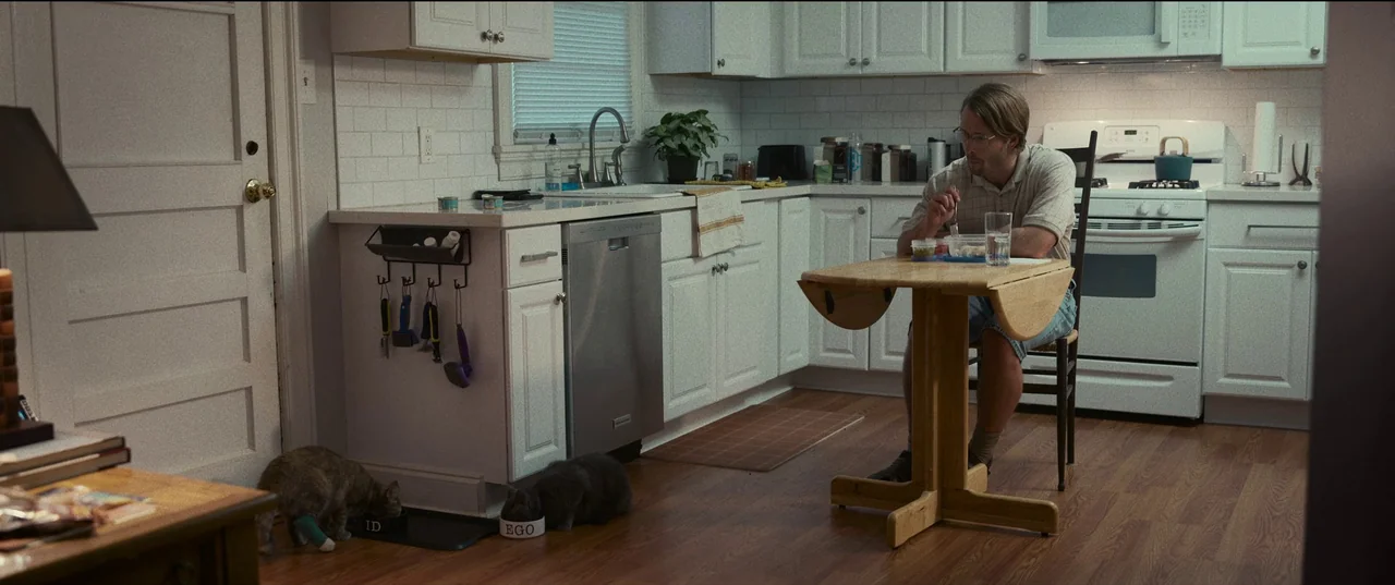 Hit Man (2023) movie still: wide — A residential kitchen features white cabinets, subway tile backsplashes, and a hardwood floor. A man sits at a…
