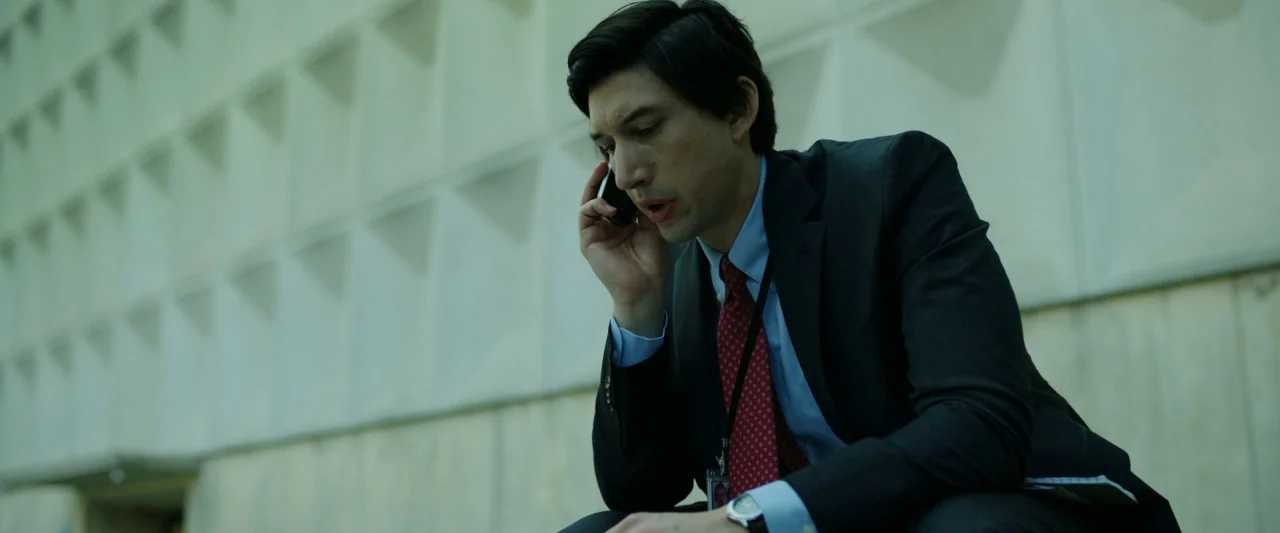 The Report (2019) movie still: medium — A man in a dark business suit sits against a textured precast concrete wall. He holds a black smartphone to…