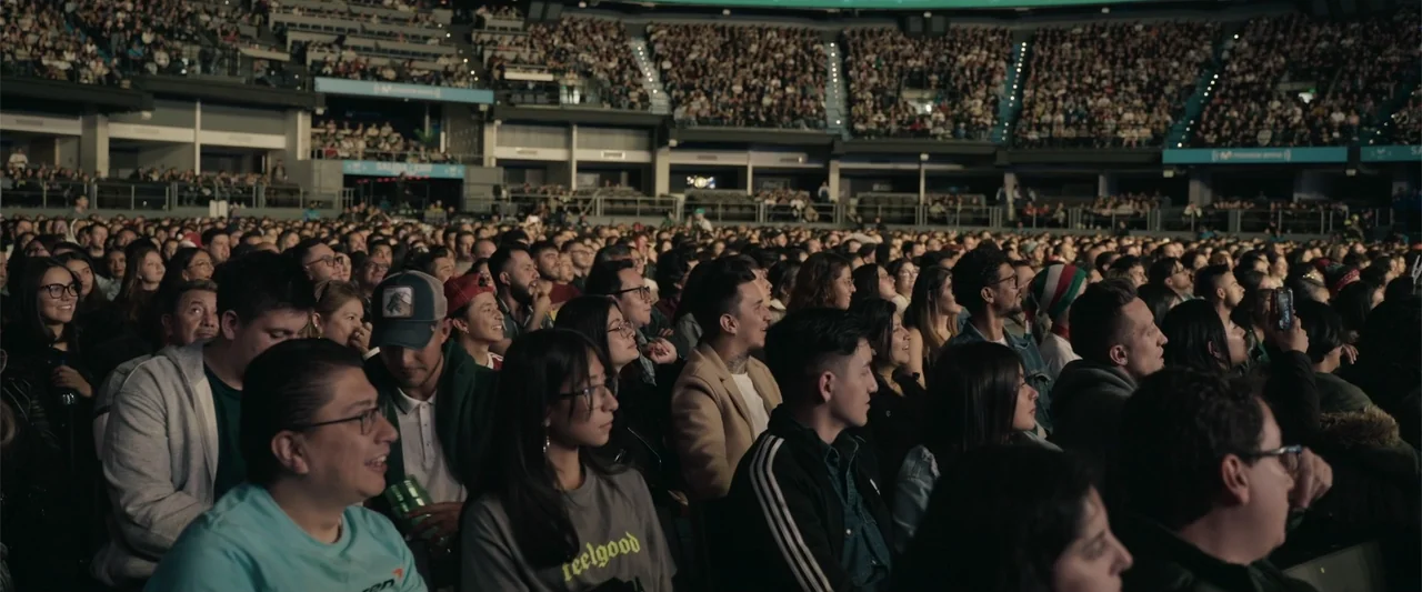 Making of: F*CKS NEWS (2025) movie still: medium — A dense crowd of people sits in rows within a darkened indoor sports arena. The audience looks…