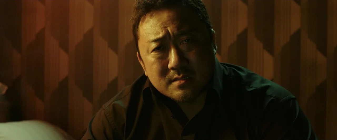 The Gangster, the Cop, the Devil (2019) movie still: closeup — A dimly lit room features a patterned wallpaper background with brown geometric…